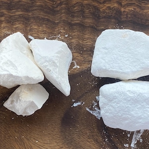 May include: Four irregular, chalky white mineral chunks with a matte, powdery texture. The pieces vary in size and shape, displaying sharp, jagged edges and a porous, opaque appearance. Some fine white dust is scattered around the base of the stones.