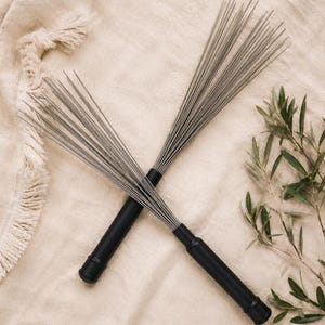 Peut inclure: Deux paires de balais de batterie avec des manches noirs et des fils fins et argentés. Les balais sont croisés sur un fond de tissu beige, avec une branche de feuilles vertes dans le coin. Les baguettes sont conçues pour la percussion.