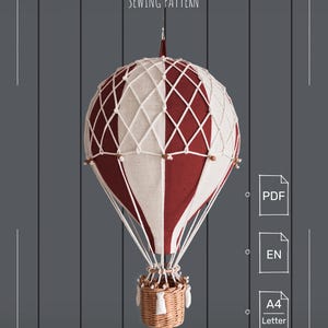 May include: A hot air balloon sewing pattern featuring a red and cream fabric balloon with a white rope net overlay and a woven wicker basket. The text reads HOT AIR BALLOON SEWING PATTERN, PDF, EN, A4, and Letter on a dark gray background.