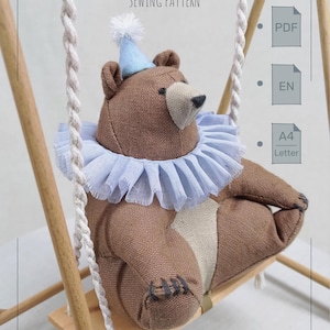 Könnte beinhalten: Ein brauner Stoffbär mit hellblauem Rüschenkragen und passendem Partyhut mit weißem Bommel, der auf einer Holzschaukel sitzt. Der Text CIRCUS BEAR SEWING PATTERN, PDF, EN und A4 Letter ist auf dem Bild deutlich zu erkennen.