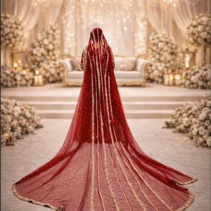 May include: A red bridal veil with gold embroidery and trim. The veil is long and flowing, with a detailed pattern. The background features a wedding ceremony setting with floral arrangements, chandeliers, and a sofa.