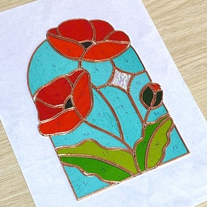Op de afbeelding: Een glas-in-lood paneel met rode klaprozen met groene bladeren tegen een turquoise achtergrond. Het ontwerp is omlijnd met koper en heeft genummerde secties. Het kunstwerk ligt op een wit oppervlak.