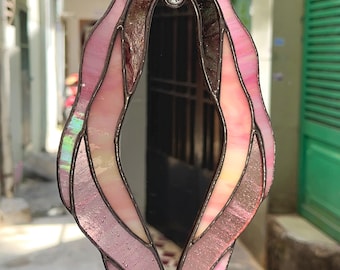May include: A stained glass suncatcher featuring an elongated, organic shape with a central mirror panel. The piece is constructed from various shades of pink and iridescent glass segments, all held together by dark metal solder lines and a small loop.