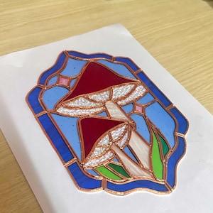 May include: A stained glass panel depicting two mushrooms with red caps and white gills, set against a light blue backdrop. The design is framed by a dark blue border with copper lines, and green leaves.