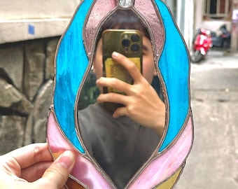 May include: A hand-held stained glass mirror with a unique, organic shape. The mirror is framed with a combination of blue, pink, and gold-colored glass pieces, creating a vibrant and artistic design. The mirror reflects the person holding it.