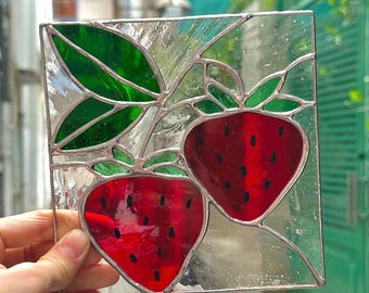 May include: A square stained glass panel featuring two red strawberries with black seeds and green leaves. The glass is clear with silver solder lines, creating a vibrant, decorative piece. The panel is held by a hand.