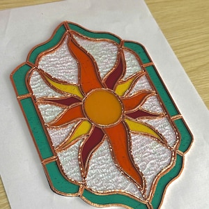 May include: A stained glass sunburst design with a copper frame. The sun features a yellow center with orange and red rays, set against a textured white background, and framed by a green border. The artwork is displayed on a white surface.