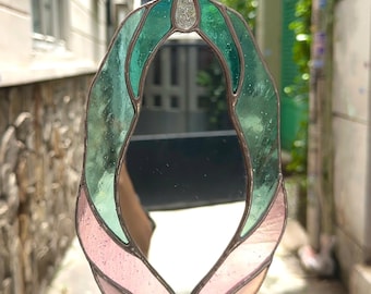 May include: Stained glass mirror in an oval shape, featuring green and pink glass panels. The mirror has a silver-colored metal frame and a small loop for hanging. The design is reminiscent of a stylized leaf or flower.
