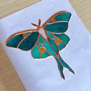 May include: A stained glass moth with copper edging. The moth features teal and green wings with yellow and white accents. The wings are outlined in copper, and the moth is set against a white background.