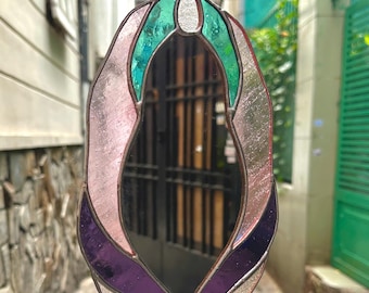 Puede incluir: Un espejo de vidrieras con una forma orgánica única. El espejo presenta paneles de vidrio rosa, turquesa, morado y transparente, enmarcados con metal plateado. El espejo está diseñado para colgarse.
