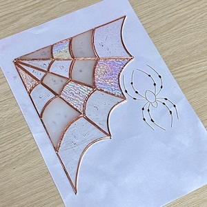 May include: A stained glass spider web design in progress, featuring iridescent and clear glass sections, copper foil, and a hand-drawn spider. The design is on white paper, with numbers on the glass.