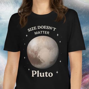 Könnte beinhalten: Schwarzes T-Shirt mit dem Text "SIZE DOESN'T MATTER" über einer Grafik von Pluto. Das Wort "Pluto" ist unter der Planetengrafik aufgedruckt. Der Hintergrund zeigt ein Galaxien-Design.