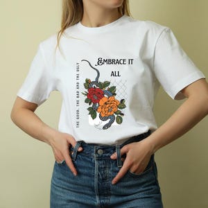 "Woman wearing white spiritual t-shirt with snake, red and orange flowers, and 'Embrace It All' quote – tattoo-style graphic design, mindful aesthetic gift for her"