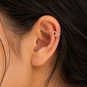 May include: A silver-toned ear cuff featuring a smooth, continuous wave-like design. The metal band curves gently along the upper cartilage of the ear, creating a simple, fluid shape that rests securely on the outer edge of the ear structure.