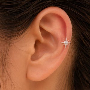 May include: A silver-toned metal ear stud featuring a North Star design with eight radiating points. The centre of the star is set with a small, clear, round gemstone, and the points are accented with tiny, clear, sparkling stones.