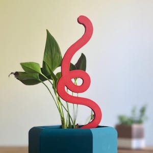Könnte beinhalten: Ein roter, stilisierter Pflanzenstecker aus Holz in Form einer Schlange, platziert in einem geometrischen Topf in Türkis und Blau. Der Stecker krümmt sich nach oben, mit einem Loch in der Nähe der Mitte. Grüne Blätterpflanzen befinden sich im Topf.