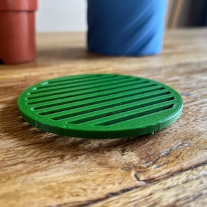 Puede incluir: Un posavasos redondo y verde para macetas con un diseño ranurado. El posavasos está hecho de un material sólido y está sobre una superficie de madera. Otras macetas son visibles en el fondo.