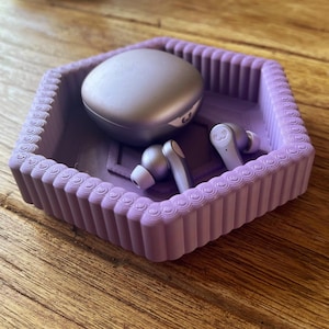 May include: A lavender hexagonal tray with a fluted edge holds a silver wireless earbud case and two earbuds. The earbuds and case are a matching silver color. The tray sits on a wooden surface.
