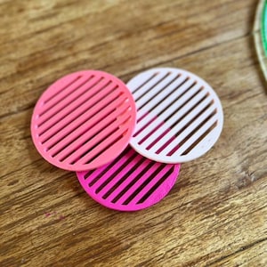 Puede incluir: Tres posavasos redondos en tonos rosa y blanco con un diseño de listones. Los posavasos están dispuestos sobre una superficie de madera. Los posavasos son un artículo decorativo.