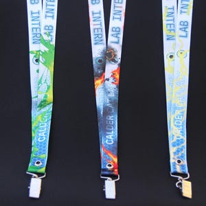 May include: Three lanyards with the text "LAB INTERN" and "CALDER CAT LABS". The lanyards have different designs: green with a splash pattern, a fiery design, and a blue and yellow lightning pattern. Each lanyard has a metal clip.