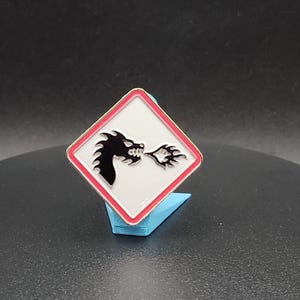 May include: A square pin with a white background, red border, and a black dragon design breathing fire. The pin is displayed on a blue stand. The dragon is facing right.