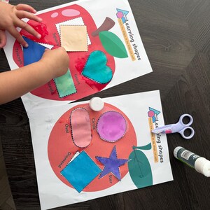 May include: Two educational sheets with apple designs for learning shapes. One shows a child matching shapes. The other has cut-out shapes. Scissors and a glue stick are also visible on a dark wooden surface.