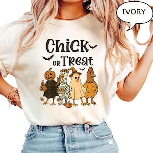 May include: Ivory-coloured t-shirt featuring the text "Chick or Treat" and a graphic of four chickens in Halloween costumes: a pumpkin, a mummy, a ghost, and a witch. The shirt is made of a soft material.