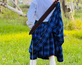 May include: A person wearing a black beret, white shirt, and a blue and black plaid kilt. A brown leather sword sheath is slung over the shoulder. White socks and black shoes complete the traditional Scottish attire, set against a grassy backdrop.