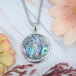 Op de afbeelding: Een zilverkleurige hangerketting met een ronde hanger met een levensboomontwerp. De hanger heeft een parelmoeren achtergrond, een zilveren boomontwerp en is omgeven door kleine, heldere kristallen. De ketting wordt gepresenteerd op een licht oppervlak met bloemen.