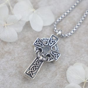 Peut inclure: Un collier pendentif en métal argenté arborant une croix celtique avec des motifs de nœuds complexes et un emblème Claddagh central. La croix est suspendue à une chaîne carrée assortie, avec le poinçon S925 visible sur le côté du pendentif.