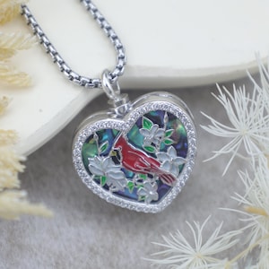 Puede incluir: Collar con colgante en forma de corazón con cadena plateada. El colgante presenta un pájaro cardenal rojo y flores blancas sobre un fondo brillante, enmarcado por un borde de pequeñas piedras transparentes. Una joya delicada.