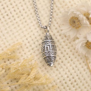 Può includere: Una collana con pendente color argento con un ciondolo a forma di barile. Il ciondolo presenta simboli e testo incisi, sospeso da una catena a maglie. Lo sfondo include fiori bianchi e piante secche.