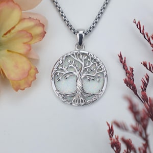 Könnte beinhalten: Eine silberfarbene Halskette mit Anhänger, die einen runden Anhänger mit einem Lebensbaum-Design zeigt. Der Baum ist vor einem weißen, opalisierenden Hintergrund platziert. Die Halskette hat eine zarte Kette und wird auf einer weißen Oberfläche mit floralen Akzenten präsentiert.