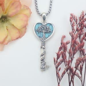 May include: A silver-toned key pendant necklace featuring a heart-shaped head with a turquoise-colored inlay and a detailed tree motif. The key shaft is decorated with a coiled snake design, and the piece hangs from a sturdy silver-toned chain.