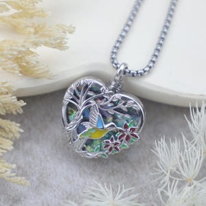 Könnte beinhalten: Halskette mit herzförmigem Anhänger und Silberkette. Der Anhänger zeigt ein Kolibri-Design mit einem Baum und Blumen. Der Kolibri hat gelbe, blaue und grüne Akzente. Der Anhänger hat einen Durchmesser von ca. 2,5 cm.