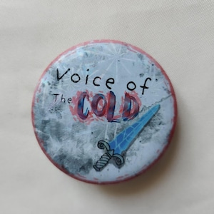 Può includere: Bottone rotondo con la scritta "Voice of the Cold" in nero e rosso. Lo sfondo è grigio ghiacciato con un'illustrazione di spada blu. Il bottone ha un bordo rosso.