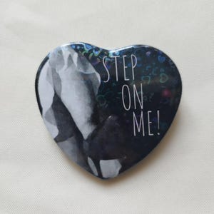 Può includere: Bottone a forma di cuore con la scritta bianca "STEP ON ME!". Il bottone presenta un'immagine in bianco e nero di gambe e piedi. Il bottone ha un bordo olografico.