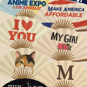 Może przedstawiać: Kolekcja dekoracyjnych wachlarzy papierowych z r&oacute;żnymi wzorami i tekstami. Niekt&oacute;re wachlarze zawierają teksty takie jak "2025 ANIME EXPO LOS ANGELES", "I ❤️ YOU" i "MAKE AMERICA AFFORDABLE". Inne wachlarze przedstawiają twarz kota, słowa "MY GIA!", literę "M" i "THE ARK KNIGHT".