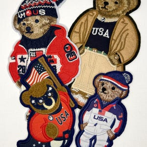 May include: Four embroidered patches featuring brown teddy bears dressed in various outfits. One bear wears a red jacket and hat with the text FAMOUS USA. Another wears a tan coat and USA shirt. A third holds a flag, and the fourth wears a white USA suit.