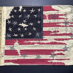 May include: A rectangular fabric flag featuring a distressed American flag design with red and off-white horizontal stripes and a dark blue canton with embroidered white stars. The left side has a tan panel with the text 67 R.L. and 77308.