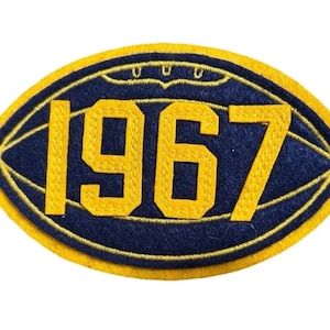 Puede incluir: Un parche bordado de forma ovalada con un diseño de balón de fútbol americano en azul oscuro y borde amarillo. Los números 1967 están bordados en una fuente grande de color amarillo en el centro, con un pequeño detalle de costura arriba.