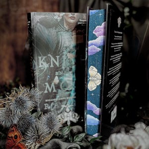 May include: Two hardcover books with decorative covers. One features a woman in armor and the title "Knight Moon." The other book has a blue spine with a butterfly and cloud design. Dried thistles and a butterfly are in the foreground.