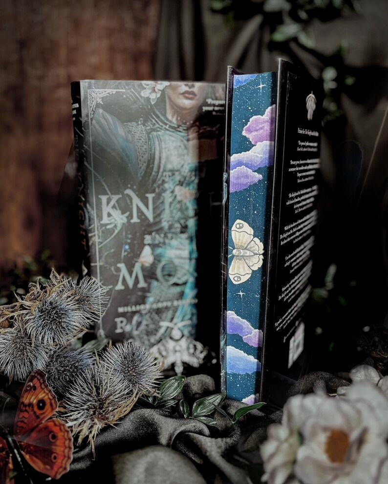 May include: Two hardcover books with decorative covers. One features a woman in armor and the title "Knight Moon." The other book has a blue spine with a butterfly and cloud design. Dried thistles and a butterfly are in the foreground.
