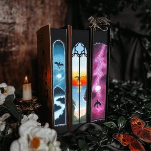 May include: A collection of fantasy books with custom painted edges depicting dragons, landscapes, and lightning. A bronze dragon bookend sits atop the books. A lit candle and decorative butterflies are in the foreground.