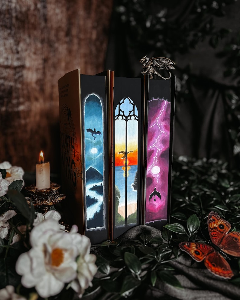 May include: A collection of fantasy books with custom painted edges depicting dragons, landscapes, and lightning. A bronze dragon bookend sits atop the books. A lit candle and decorative butterflies are in the foreground.