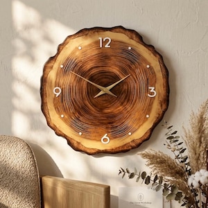May include: A round wall clock crafted from a natural wood slice featuring visible dark brown tree rings. The clock face displays white numerals at the 12, 3, 6, and 9 o'clock positions, with small white dots for other markers and thin gold-toned metal hands.