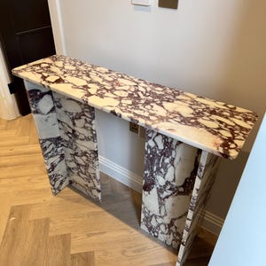 May include: A rectangular marble console table with a white and burgundy pattern. The table has a long, flat top and two supporting legs. The legs are also made of marble and have a similar pattern. The table is placed against a light-colored wall.