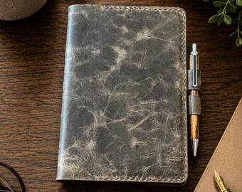 May include: A rectangular notebook with a distressed dark gray leather cover featuring a marbled texture and visible stitching along the edges. A wooden pen is held in a matching leather loop attached to the side of the notebook cover.