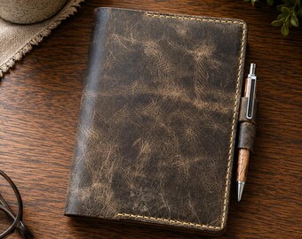 May include: A dark brown leather notebook cover with a distressed, textured finish and visible yellow stitching along the edges. A wooden pen is held in a matching leather loop attached to the side of the cover, completing the set.