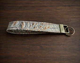 May include: A wristlet keychain featuring a textured, light gray and tan faux-alligator skin pattern with visible brown stitching along the edges. The strap is secured by a rectangular antique brass-toned metal clasp attached to a circular ring.